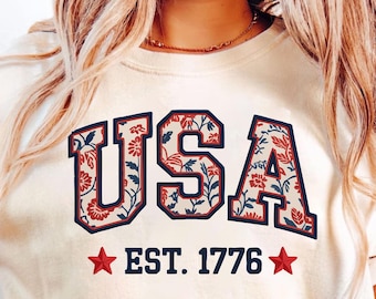 May include: Cream-colored t-shirt with the word "USA" in a navy blue, floral-filled font. Below the text are two red stars and the text "EST. 1776". The shirt has a relaxed fit and short sleeves.