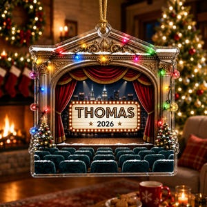 May include: A clear, flat, theater-themed Christmas ornament featuring a stage with red curtains, rows of dark seats, and a marquee sign that reads THOMAS 2026. The frame is decorated with gold architectural columns and colorful string lights.