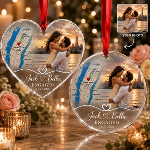 May include: Two heart-shaped glass ornaments featuring a printed map of New York City with a red heart icon, a photograph of a couple embracing by the water, and the text "Jack & Bella ENGAGED 11.23.2026" suspended by red satin ribbons.