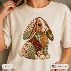 May include: An ivory short-sleeve t-shirt featuring a large graphic of a rabbit constructed from various patchwork fabric patterns, including floral, plaid, gingham, and polka dot designs in shades of brown, green, red, and cream.