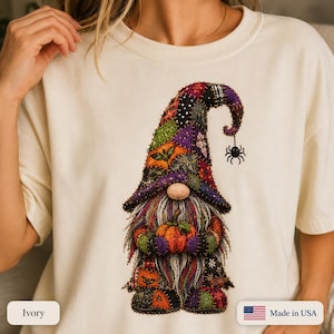 May include: An ivory t-shirt featuring a graphic of a gnome with a patchwork-style hat and clothing in shades of purple, orange, green, and black. The gnome has a long gray beard, holds a small orange pumpkin, and has a black spider hanging from its hat.