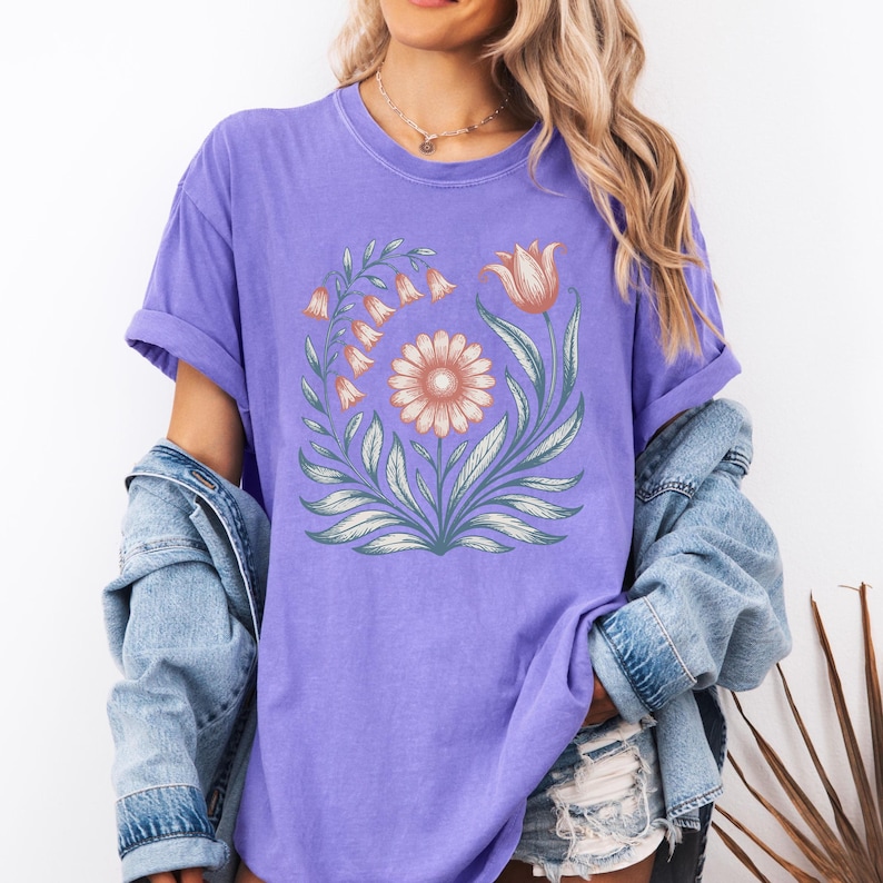 May include: A lilac-colored t-shirt featuring a floral design. The design includes a central daisy, a tulip, and bell-shaped flowers, all in soft peach and orange hues, surrounded by green leaves. The shirt has rolled-up sleeves.