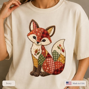 May include: An ivory t-shirt featuring a central graphic of a fox designed with a patchwork quilt aesthetic. The fox illustration incorporates various patterns including red gingham, floral prints, and green checks, outlined with visible stitching.