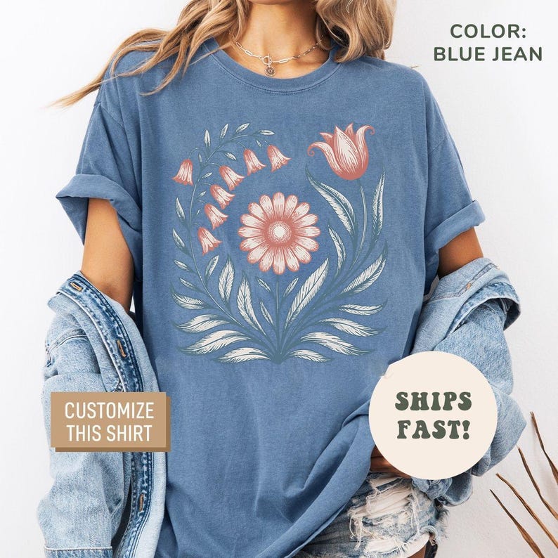 May include: Blue jean colored t-shirt featuring a floral design with a central daisy, a tulip, and bell-shaped flowers. The shirt has the text "Customize This Shirt" and "Ships Fast!"