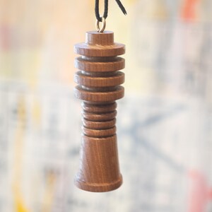 May include: A handcrafted wooden pull with a dark brown finish. The pull is cylindrical with a series of stacked, circular tiers. A black cord is attached to the top with a gold-colored ring. The background is blurred.