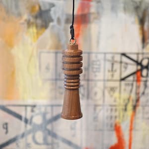 May include: A wooden pendulum with a dark cord, suspended against a colorful, abstract background. The pendulum is a light brown color, with a cylindrical top and a flared base. The design features stacked, ridged sections.