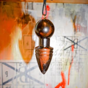 Puede incluir: Un péndulo de madera marrón oscuro con una parte superior redondeada y una base puntiaguda. Está suspendido por un cordón negro y un pequeño anillo dorado. El fondo presenta una mezcla de colores y patrones.
