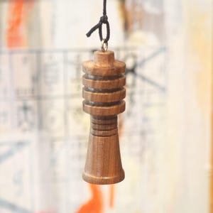 May include: A small, brown wooden object with a cylindrical shape and multiple stacked rings. It is suspended by a black string tied in a knot. The object has a smooth, polished finish and appears to be a decorative item or pendant.