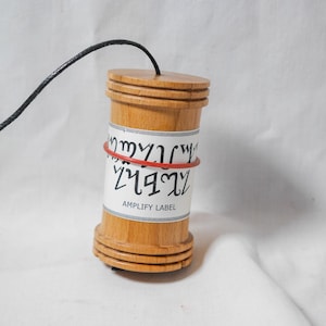 May include: A wooden cylindrical object with a black cord attached to the top. The object has a white label with text and is secured with a red rubber band. The label reads "AMPLIFY LABEL".
