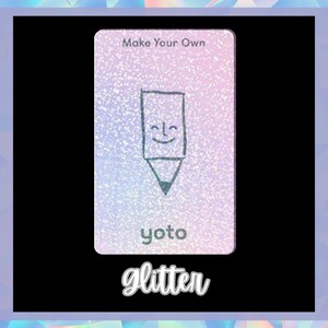 Op de afbeelding: Een witte kaart met een glinsterende textuur en een tekening van een lachend potlood. De kaart heeft bovenaan de tekst "Make Your Own" en onderaan "yoto". De tekst "glitter" staat onder de kaart.
