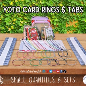 K&ouml;nnte beinhalten: Eine Auswahl an YOTO-Kartenringen und -Tabs mit dem Text "YOTO CARD RINGS & TABS" oben. Das Bild zeigt Kartenringe in verschiedenen Farben, Kartenhalter und kleine B&uuml;cher. Der Text "SMALL QUANTITIES & SETS" befindet sich unten.