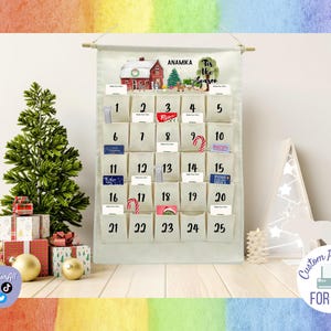 Op de afbeelding: Een witte canvas adventskalender met 25 genummerde vakjes. De kalender heeft een aquarel illustratie van een huis met een kerstboom en de tekst "Tis the Season". De naam "Anamika" staat bovenaan de kalender gedrukt.