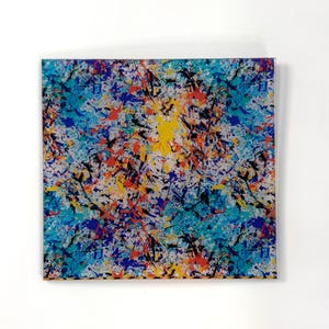May include: Square artwork with a colorful abstract splatter design. The artwork features a white background with splatters of blue, yellow, orange, and black paint. The artwork is square and appears to be on a clear surface.