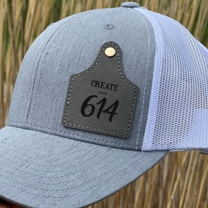 May include: A gray and white trucker hat with a leather patch that says "CREATE with 614".