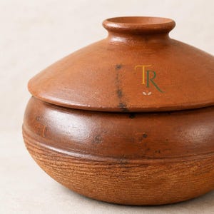 Peut inclure: Un pot en terre cuite de couleur ocre avec un couvercle assorti. Le pot présente une partie supérieure lisse et une base texturée et nervurée. Le couvercle possède un bouton central et affiche les lettres TR en vert et jaune avec une petite feuille.