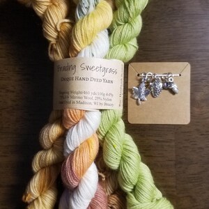 May include: Four skeins of hand-dyed yarn in shades of yellow, green, orange, and white. The yarn is labeled "Brading Sweetgrass Unique Hand Dyed Yarn" with the details "Fingering Weight/460 yds/100g 4-Ply 75% SW Merino Wool, 25% Nylon Dyed in Madison, WI by Breezy".