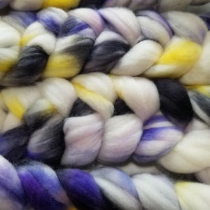 May include: Close-up of a braided roving in shades of white, yellow, purple, and black. The thick wool fiber is arranged in a textured pattern, ideal for fiber art projects. The color transitions create a visually appealing effect.