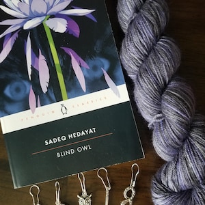 May include: A book titled "Blind Owl" by Sadeq Hedayat, a Penguin Classics edition, with a purple and gray yarn skein and five silver stitch markers. The stitch markers include a small owl, a snake, a needle, a teardrop-shaped crystal, and a black bead with an evil eye charm.