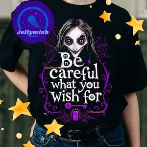 May include: Black t-shirt featuring a graphic of a character with button eyes and the text "Be careful what you wish for" in white and purple. The design includes a key and spiderwebs.