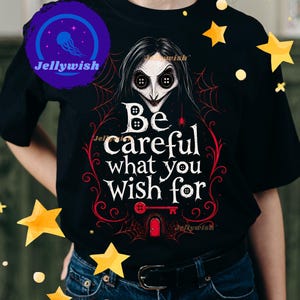 May include: Black t-shirt featuring a stylized illustration of a character with button eyes and the text "Be careful what you wish for." The design includes a spiderweb, key, and door in red and white.