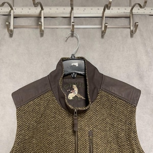 May include: A brown and green herringbone vest with a brown leather collar and shoulder accents. The vest has a full zipper and a small zippered pocket. The brand "ORVIS" is embroidered on the chest, along with a bird emblem.