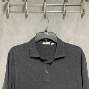 May include: A dark gray polo shirt featuring a classic collar, a short button placket with two visible buttons, and long sleeves. The shirt is displayed on a black plastic hanger, showcasing a textured fabric construction and a simple design.