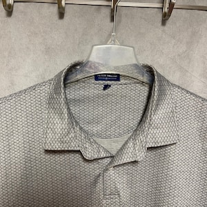 以下が含まれることがあります： 透明なプラスチックハンガーに掛けられた、テクスチャード加工のパターンが施されたライトグレーのポロシャツ。 シャツは襟付きで、ボタン付きの前立てが付いています。 ブランド名「PETER MILLAR」が小さなタグに表示されています。