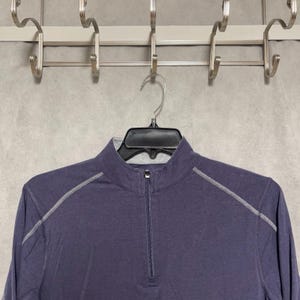 May include: A long-sleeved, navy blue athletic shirt with a quarter-zip closure. The shirt features a mock neck and light gray stitching along the shoulders and sleeves. The shirt is hanging on a black hanger.