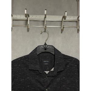 May include: A black button-down shirt with white flecks, hanging on a black hanger. The shirt has a collared neckline and the brand name "BUGATCHI" is visible on a tag. The shirt is hanging on a metal rack.
