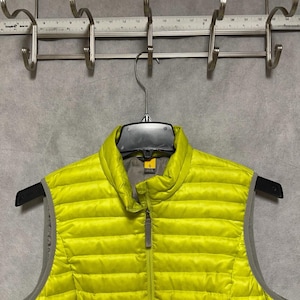 May include: A bright yellow puffer vest with a full-length zipper and a gray interior. The vest is hanging on a black hanger against a gray background. The vest has a high collar and a small logo on the chest.