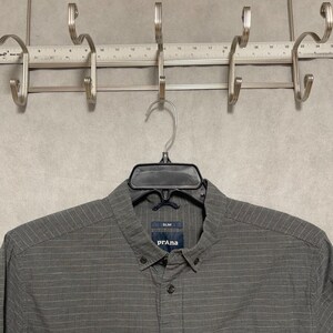 Könnte beinhalten: Ein graues Button-Down-Hemd mit dem Markennamen "prAna" auf dem Etikett. Das Hemd hängt an einem schwarzen Kleiderbügel. Das Hemd hat ein strukturiertes Muster und einen Button-Down-Kragen.