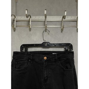 Pode incluir: Close-up de jeans pretos pendurados em um cabide preto. Os jeans têm um design clássico de cinco bolsos e um fecho de botão. Os jeans estão pendurados em um rack de metal com ganchos.