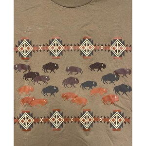 May include: A tan t-shirt featuring a repeating pattern of bison and geometric designs in shades of brown, red, and black. The bison are depicted in various colors, creating a visually appealing design. The shirt has a crew neck.