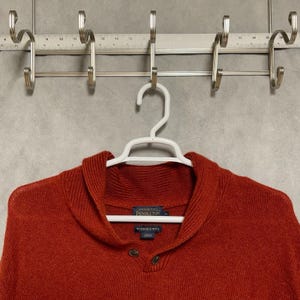 May include: A rust-coloured sweater with a shawl collar hangs on a white hanger. The sweater has a buttoned placket and a label that reads "PENDLETON WASHABLE WOOL". The hanger is suspended from a metal rack with multiple hooks.