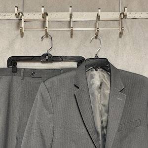 May include: A grey suit consisting of a blazer and trousers with a subtle herringbone pattern. The garments are hung on black plastic hangers from a silver metal rack that includes a ruler showing measurements in inches along the top edge.