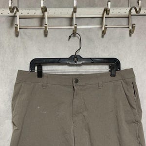 May include: Taupe-colored trousers featuring a textured fabric, a central button closure, belt loops, and a zippered pocket on the right side. The trousers are suspended from a black plastic hanger against a wall-mounted metal rack.