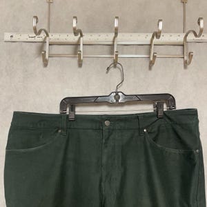 Op de afbeelding: Donkergroene broek hangend aan een zwarte hanger. De broek heeft twee voorzakken en een knoopsluiting. Boven de broek bevindt zich een metalen rek met haken.