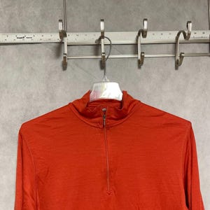 May include: A long-sleeve, quarter-zip pullover shirt in a solid burnt orange color. The garment features a high collar with a vertical zipper closure and visible stitching along the seams, hanging from a clear plastic hanger.