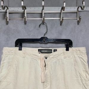 May include: Beige Old Navy pants hanging on a black hanger. The pants have a drawstring waist and a button closure. The Old Navy label is visible. The pants are hanging in front of a gray background with a metal rack.