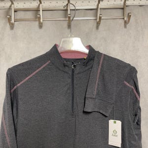 May include: A charcoal gray long-sleeve shirt with a quarter-zip neckline. The shirt features pink stitching along the seams and a tag with the brand name "tasc". The shirt is hanging from a metal hanger.