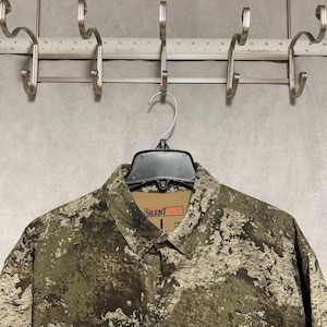Puede incluir: Camisa con botones de camuflaje con la palabra "SILENT" en la etiqueta. La camisa es principalmente verde y marrón con un patrón moteado. Cuelga de una percha negra en un perchero de metal.