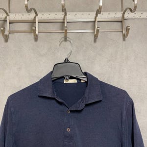 May include: A navy blue long-sleeve polo shirt with a button-down collar. The shirt is hanging on a black hanger. The shirt has three buttons down the front.
