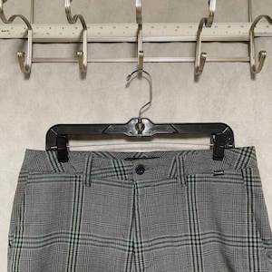May include: Gray and black plaid pants hanging on a black hanger. The pants have a classic plaid pattern with a button and zipper closure. The pants are hanging on a metal rack with hooks.