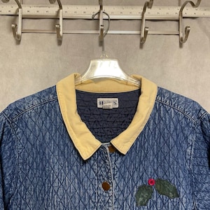 May include: A blue quilted jacket with a tan corduroy collar and button closure. The jacket features a diamond pattern and a small patch with holly leaves and berries. The jacket is hanging on a metal hanger.