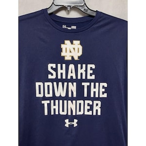 Könnte beinhalten: Marineblaues Sport-T-Shirt mit dem Text "SHAKE DOWN THE THUNDER" in weißen Blockbuchstaben. Das Shirt zeigt das Notre Dame-Logo in Gold und das Under Armour-Logo. Das Shirt hängt an einem schwarzen Kleiderbügel.