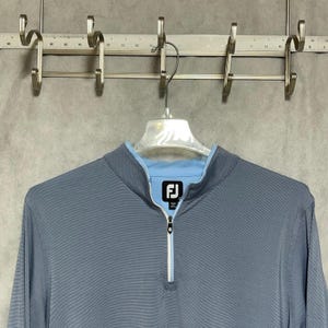 May include: A slate blue long-sleeve quarter-zip pullover featuring a light blue inner collar lining and a white zipper track. The garment displays a black and white FJ logo patch on the inner neck label, indicating the brand FootJoy.
