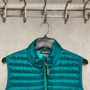May include: Teal puffer vest with a full-length zipper and a stand-up collar. The vest is hanging on a black hanger. The Eddie Bauer logo is visible on the inside of the vest.