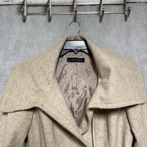 Puede incluir: Un abrigo beige de Zara Basic con cuello ancho y cierre de cremallera. El abrigo está colgado de una percha metálica. El forro interior es de color marrón claro.