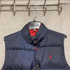 May include: Navy blue reversible puffer vest with a red interior. The vest features a high collar, snap closures, and a small red logo on the chest. The vest is hanging on a metal hanger.
