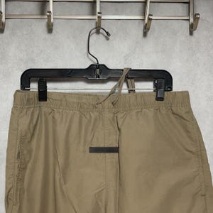 Op de afbeelding: Lichtbruine joggingbroek hangt aan een zwarte hanger. De broek heeft een donkere rechthoekige patch in het midden en een trekkoord in de taille. De broek is gemaakt van licht materiaal.